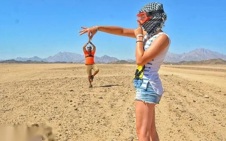 Woman wearing a snorkel mask on her head watches a man doing a jump pose in the flat desert with mountains in the background.
