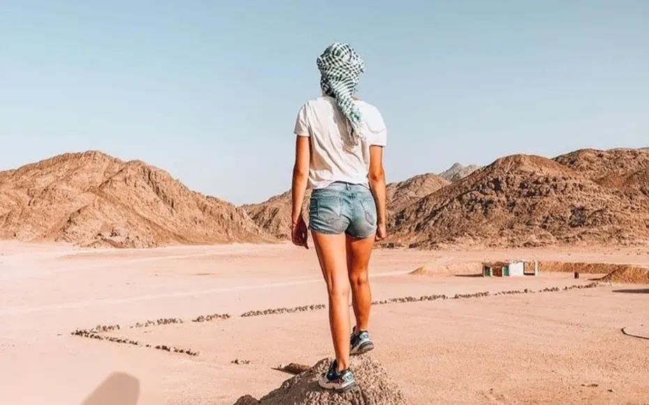 Woman wearing shorts and a headscarf walking away from the camera across flat desert ground with rocky mountains on either side.