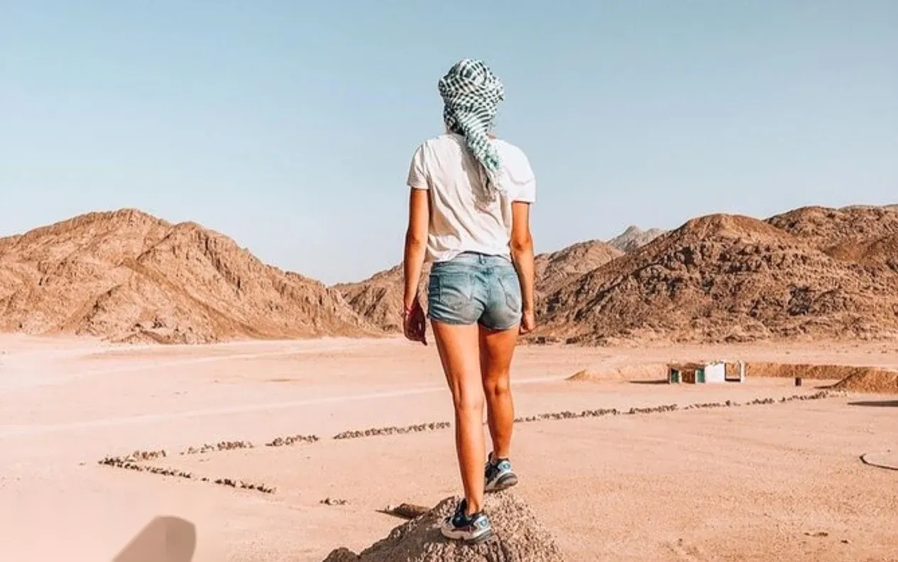 Rider wearing a checkered headscarf raises a peace sign while sitting on a quad bike among a line of other quad bikes at a desert camp.