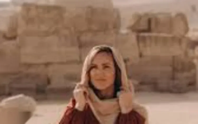 Blurry close-up portrait of a woman in a headscarf with a pyramid's stone blocks out of focus behind her.