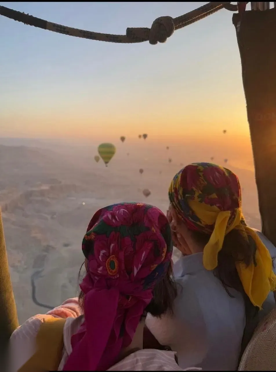 Dozens of hot air balloons scattered across the sky over the Luxor valley at sunrise, seen from inside another balloon's basket.