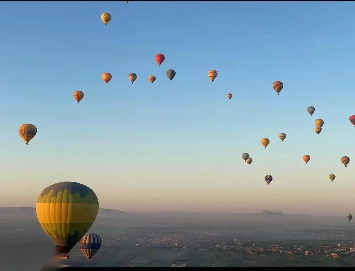 Two passengers in colorful patterned headscarves looking out from a hot air balloon basket at other balloons and the valley below.