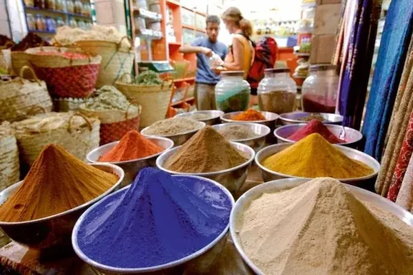 A market spice stall with pyramid-shaped mounds of colorful ground spices in bowls, tourists browsing baskets behind.