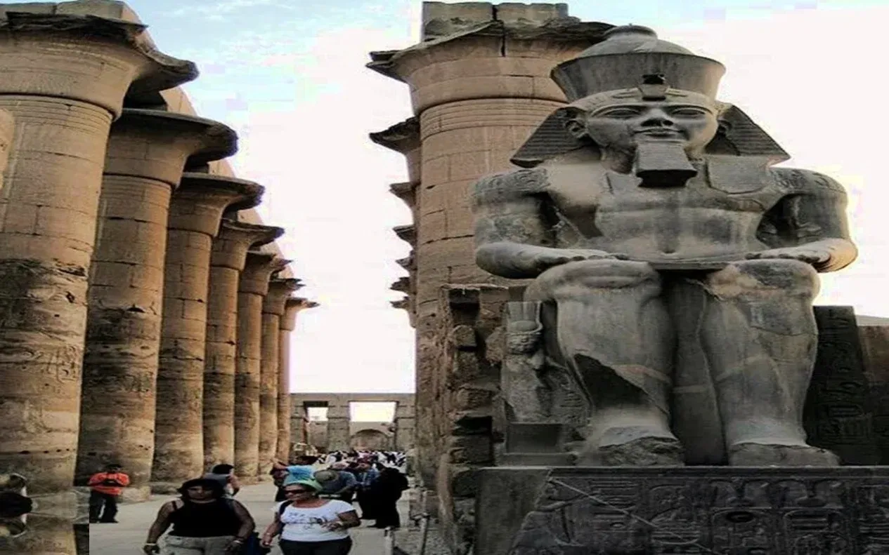 Crowd of tourists walking through Karnak Temple's Great Court among sandstone columns and ruined pylon towers, palm trees in the background under a bright blue sky.