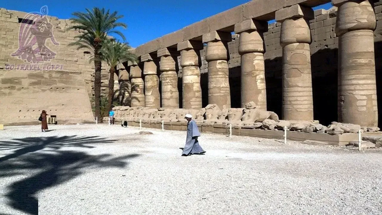 Tourists walk toward Luxor Temple's first pylon, with the single remaining obelisk and colossal statues framed between the towering stone gateways.