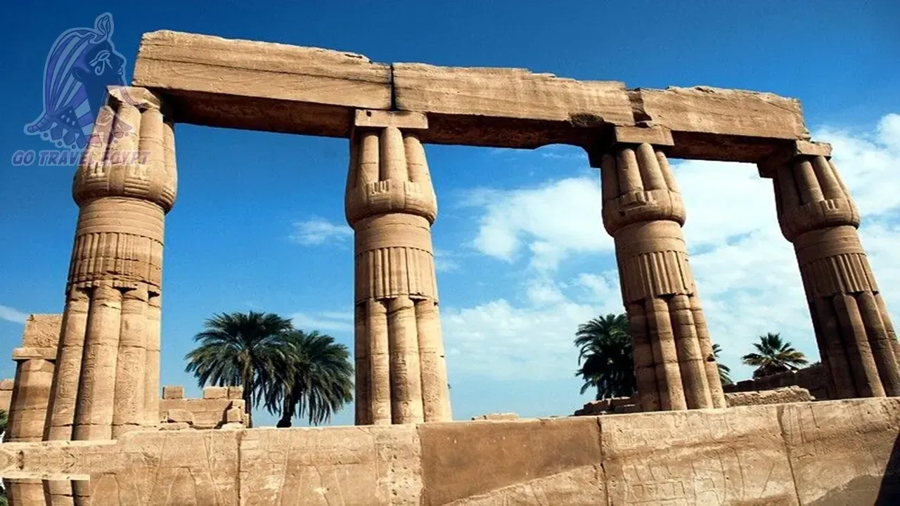 A line of visitors walks past a round stone column and a weathered sphinx statue toward the ruined pylon towers of Karnak Temple.