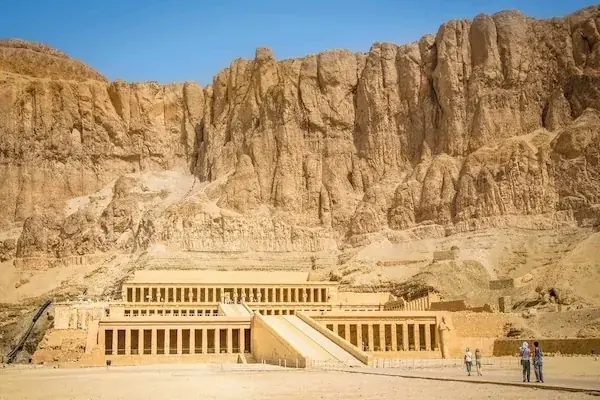 Hatshepsut Temple's terraced colonnades stand at the base of sheer desert cliffs, with a few visitors near the entrance.
