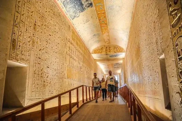Tourists walk along a wooden walkway through a long tomb corridor decorated with rows of hieroglyphic text and painted figures.