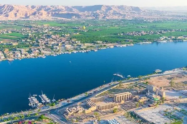 Aerial view of Luxor Temple beside the Nile River, surrounded by the city of Luxor with mountains in the distance.