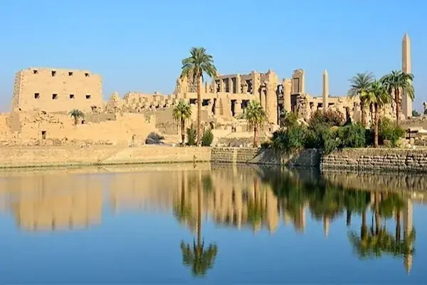 Karnak Temple's sandstone ruins, palm trees, and obelisks reflect perfectly in the still water of the sacred lake.
