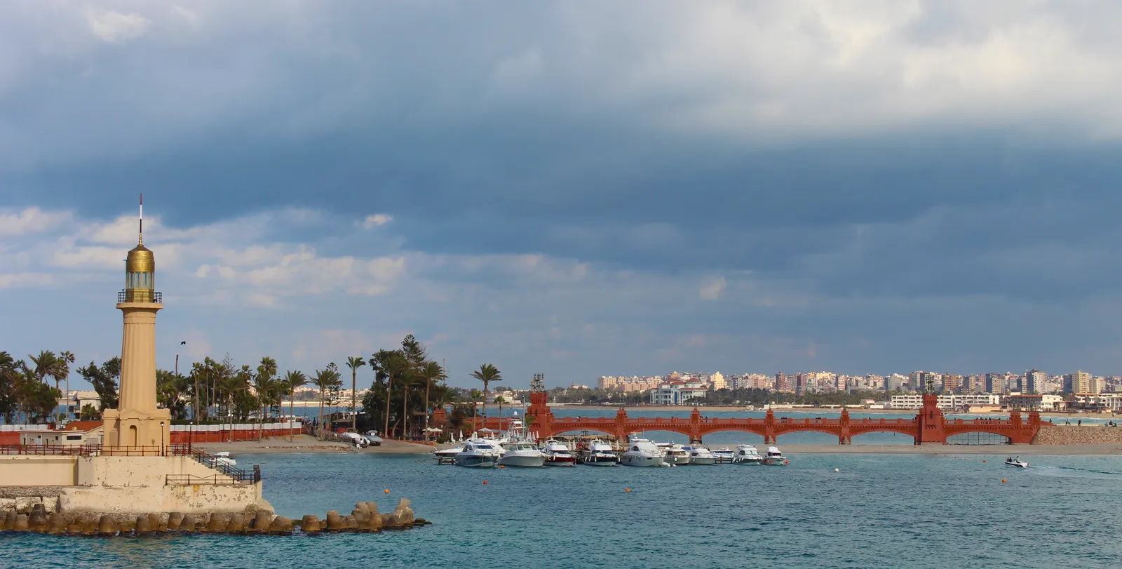 A small lighthouse stands at a pier beside a marina full of boats, a red arched bridge, and the Alexandria coastline under a cloudy sky.
