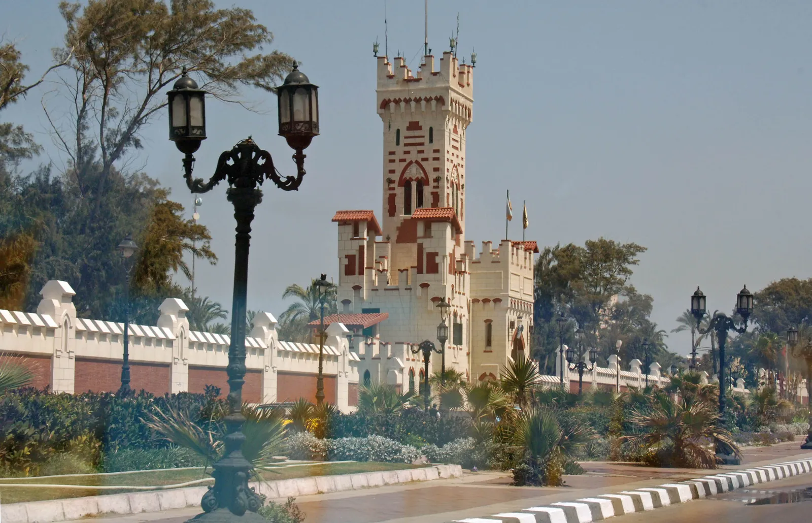 An ornate castle-style tower entrance building of Montaza Palace grounds, with wrought-iron lampposts and a white boundary wall along a tree-lined road.