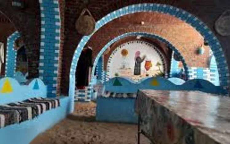The interior of a Nubian house features brick archways painted blue and white with a colorful folk-art mural of a woman, low bench seating along the walls.