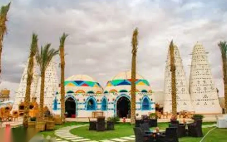 Domed Nubian-style buildings painted with colorful geometric patterns stand beside tall cone-shaped pigeon towers and palm trees under a cloudy sky.