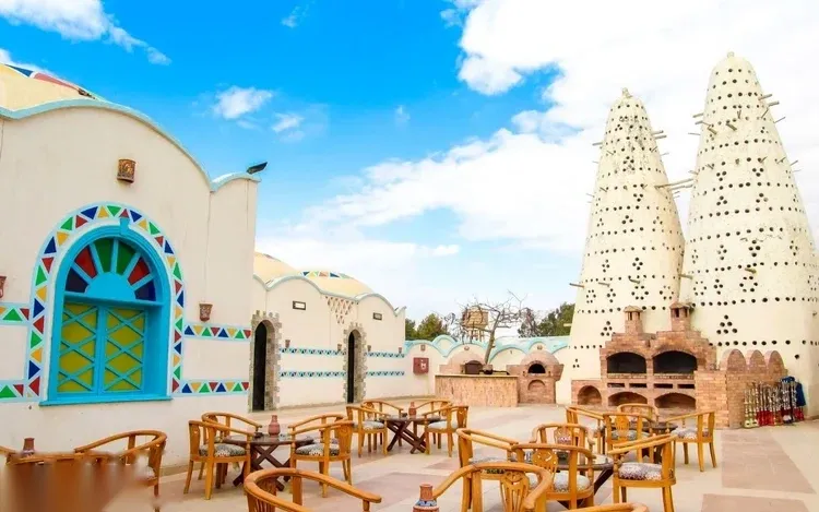 A courtyard with wooden tables and chairs sits beside a blue-and-white Nubian building with a mosaic-tiled archway and tall pigeon towers, under a partly cloudy sky.