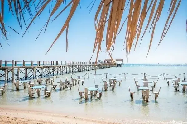 A wooden pier lined with submerged tables and chairs extends into the clear water at Orange Bay, framed by hanging palm fronds.
