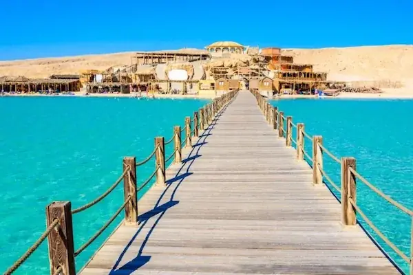 A long wooden jetty stretches from a cluster of beach huts across turquoise water toward the camera under a clear sky.