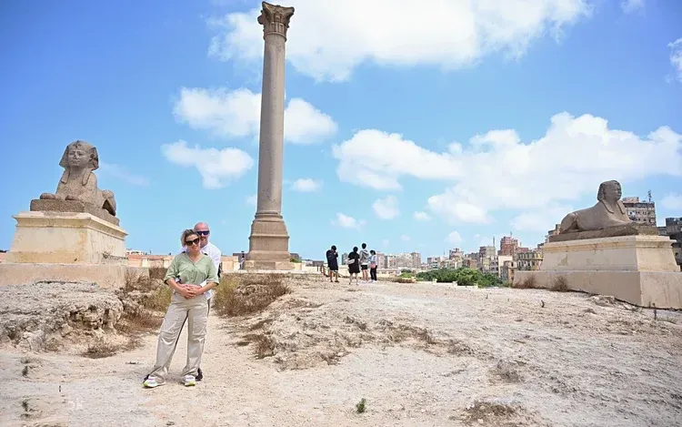 A couple stands among carved sphinx statues and a tall granite column at an ancient ruins site under a blue sky.