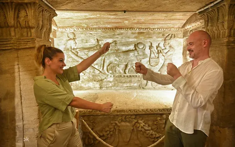 A couple play-fights with raised fists in front of an ancient carved stone relief showing Egyptian deities, inside a torch-lit catacomb chamber.