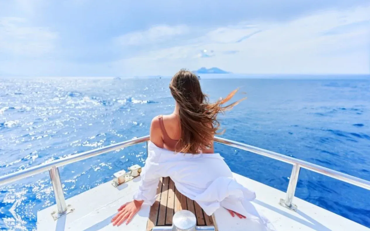 A woman in a white dress sits at the bow of a moving yacht, hair blowing in the wind, looking out at the open sea with a distant island on the horizon.