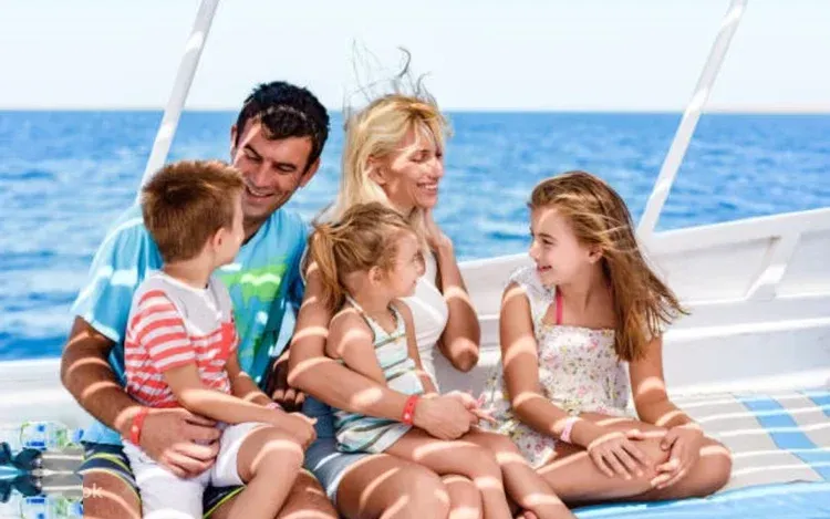 A family of two adults and three children sit together laughing on the deck of a sailing yacht at sea.