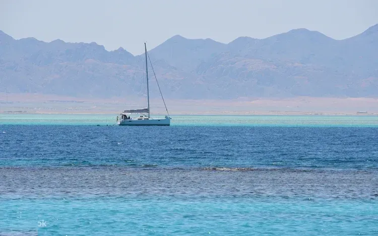 A white sailboat is anchored near a reef with desert mountains along the coastline in the background.