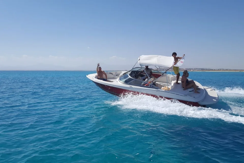 A speedboat full of passengers, some standing, races across blue water leaving a white wake, coastline visible in the distance.