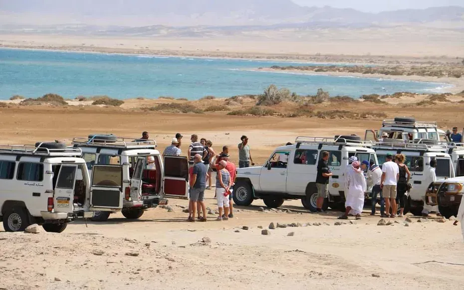 Group of tourists sitting on the ground around a campfire between two 4x4 vehicles under a tree at dusk.