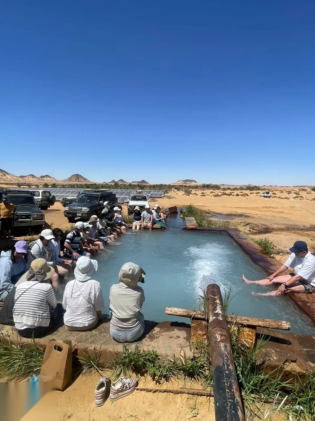 Group of tourists sitting along the edge of a warm turquoise desert spring with their feet in the water, jeeps and solar panels behind them.