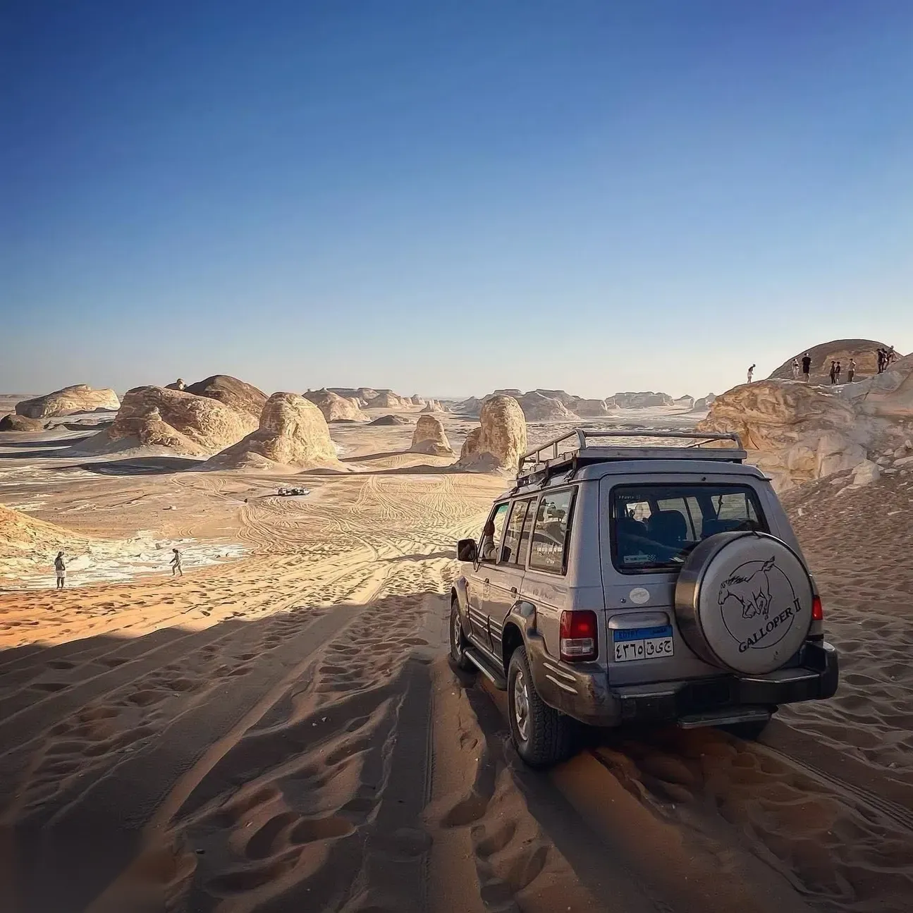 Grey 4x4 with a spare tire on the back parked overlooking a valley of white rock formations, with tourists walking below.