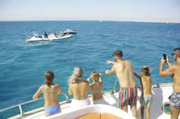 Black-hulled yacht underway at sea with passengers relaxing on the upper sun deck and coastline in the distance.