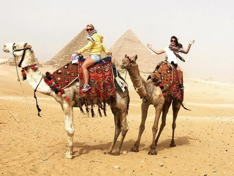 Two women riding camels side by side with a pyramid in the background, one waving to the camera.