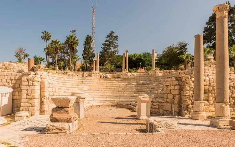 Roman amphitheater with tiered white marble seating and stone columns surrounded by palm trees.