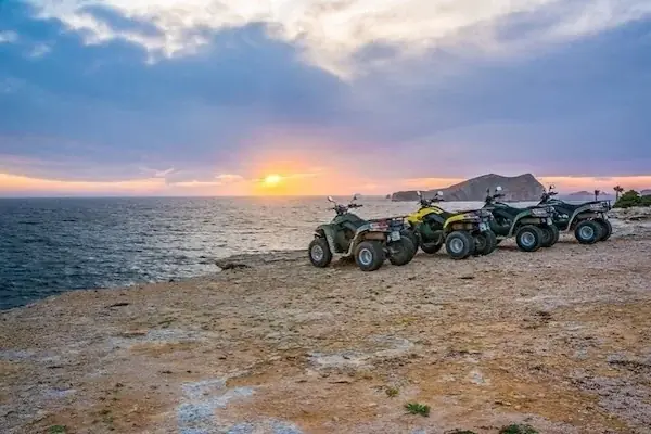 Two women wearing headscarves and sunglasses ride a red quad bike at the front of a desert convoy, with mountains in the background.