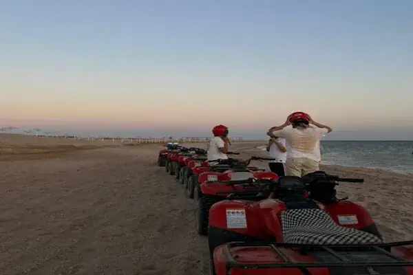 Quad bikers seen from behind riding away across flat desert sand toward distant mountains.