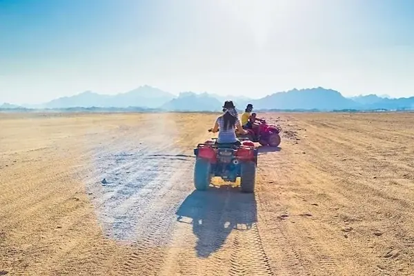 A couple sits together on a red quad bike in warm desert light, with another pair on a quad bike behind them.