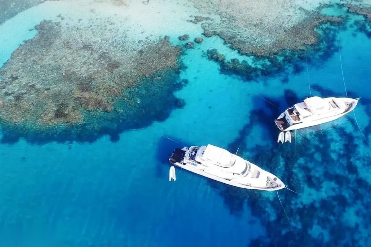 Elevated view of a curved turquoise bay with a rocky headland, sandy beach, and small boats moored in the distance.