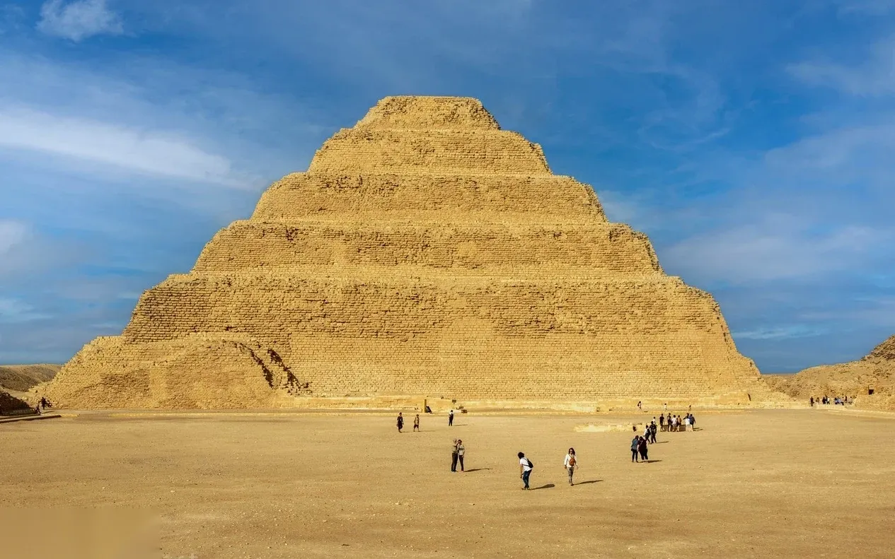 The Step Pyramid of Djoser seen up close, with visitors walking across the sand in front of its tiered stone structure.