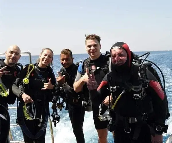 A group of five divers pose for a photo on the back of a boat, giving thumbs-up and hand signals.