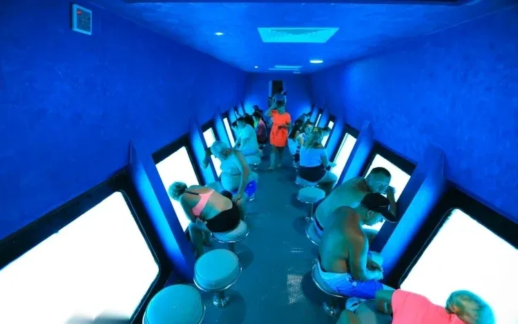 Interior of the semi-submarine's underwater viewing gallery with tourists seated on stools photographing glowing blue windows