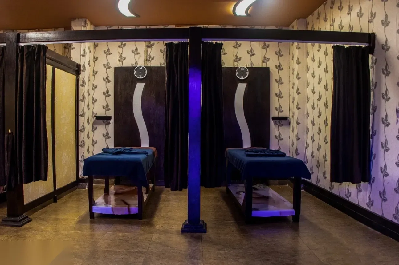 Dimly lit hammam treatment room with two massage tables facing each other beneath a purple LED accent light and curtained alcoves