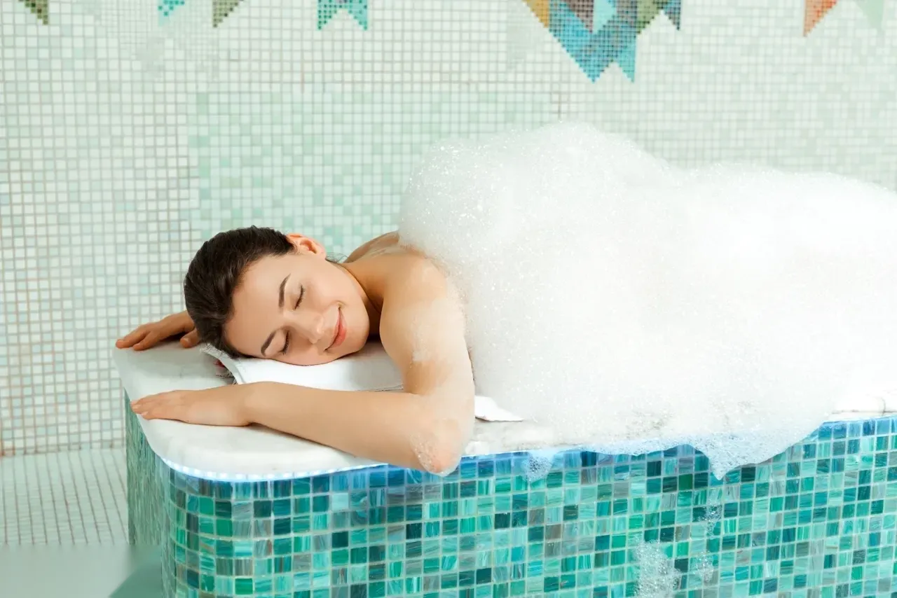 Woman wrapped in a white towel with a towel turban, reclining on a heated marble bench beside a carved stone basin under a classical relief carving