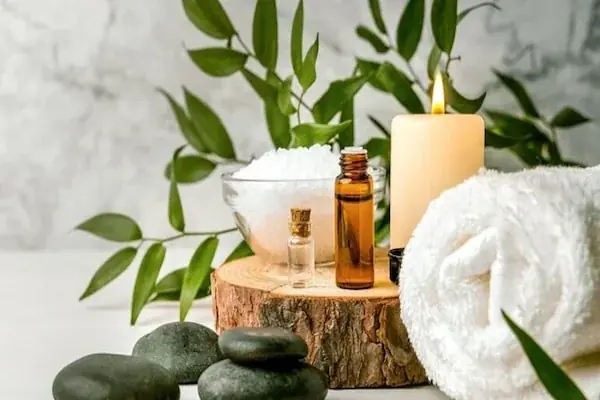 Spa flatlay on a wooden stump with essential oil bottles, sea salt, a lit candle, rolled white towel, and green foliage