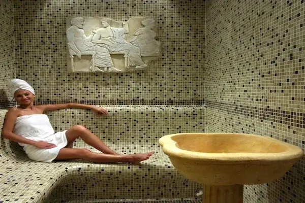 Woman wrapped in a white towel with a towel turban, reclining on a heated marble bench beside a carved stone basin under a classical relief carving