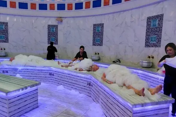 Several guests lying face-down covered in white foam on a marble hammam platform while staff attend to them under a colorful domed ceiling