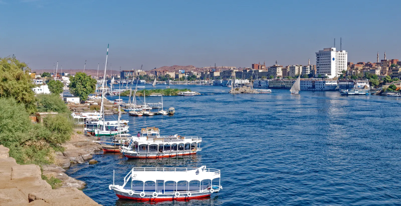 Feluccas moored at a stone riverbank dock in Aswan with city buildings behind