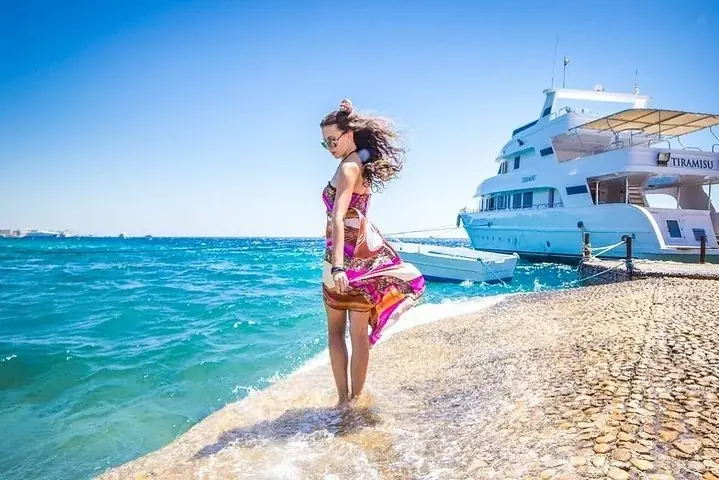 Woman in a flowing colorful dress walking along a stone jetty beside a docked yacht named Tiramisu