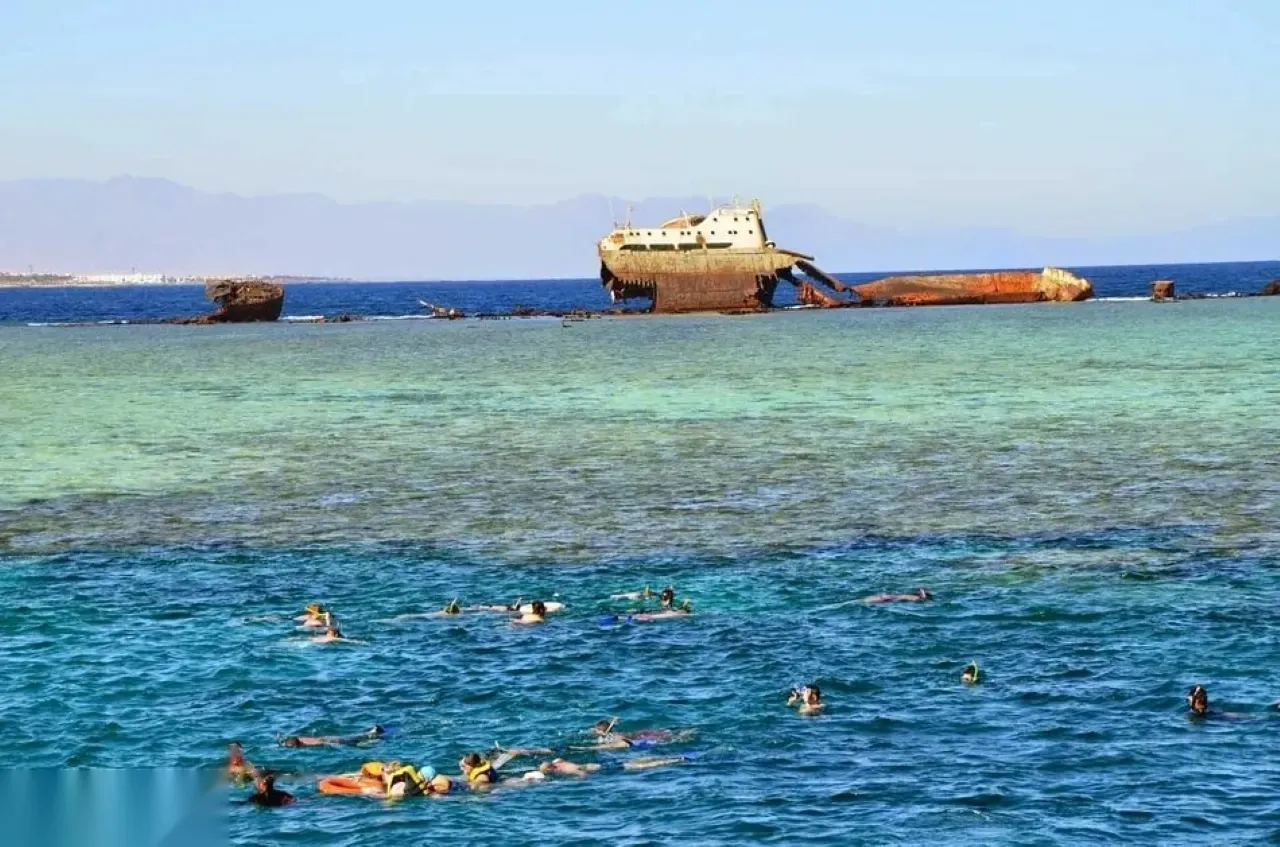 Snorkelers swimming near a reef with the rusted broken hull of a shipwreck visible on the horizon