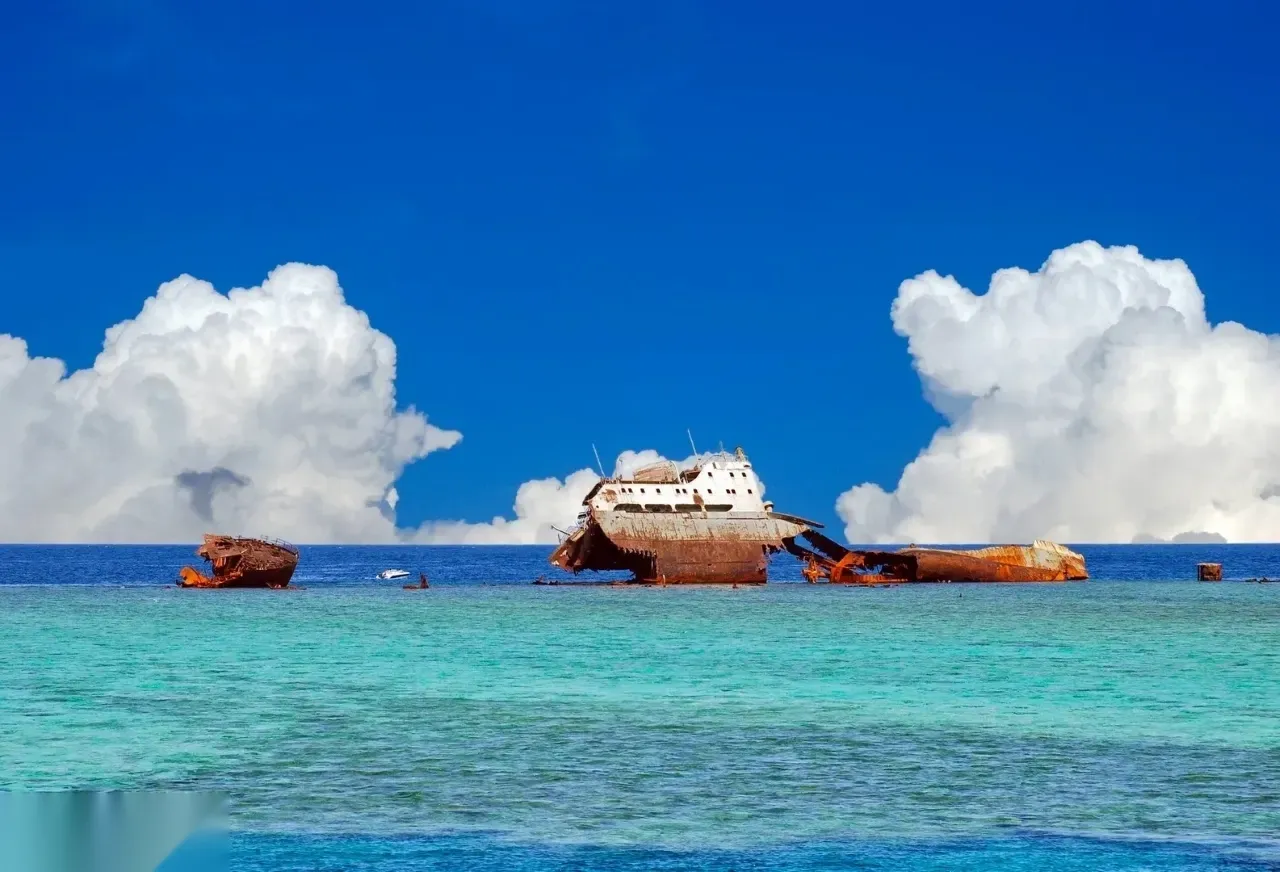 Large rusted cargo shipwreck broken apart on a reef under a dramatic sky with towering cumulus clouds