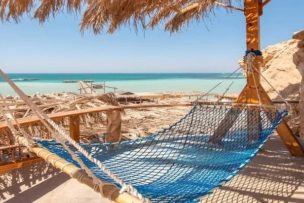 Blue rope-net hammock strung under a straw canopy overlooking a turquoise bay with a wooden jetty and distant beach huts